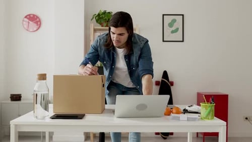 Man Writes on Box While Using Laptop at Desk