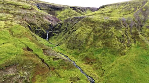 Aerial drone view of a lush green canyon where a narrow stream flows toward a secluded waterfall bet