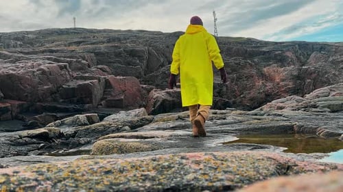 A Man Who Looks Like a Fisherman Wearing a Yellow Raincoat and a Hat Walks Along the Rocky Coast