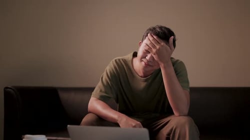 Der Mann sitzt alleine auf einem Sofa, berührt seine Stirn mit der Hand und fühlt sich müde vom Licht