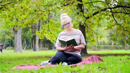 Portrait Happy Senior Woman Sitting in the Park on the Lawn Reading a Book holding in hands. Mature