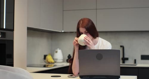 Woman Works on Laptop and Drinks in Kitchen