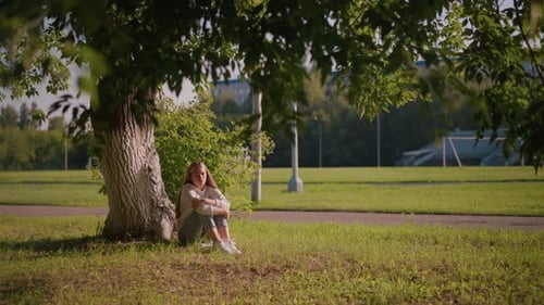 Woman Sitting Thoughtfully Under Tree with Sunlight and Stadium in Background
