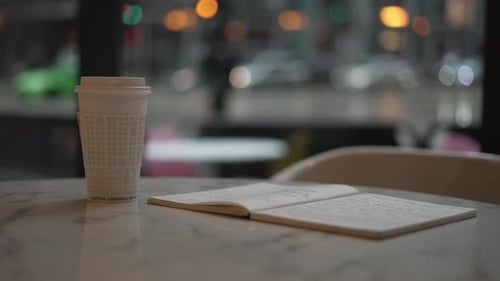 Coffee cup and open notebook on café table near window with city traffic in soft focus. Urban worksp