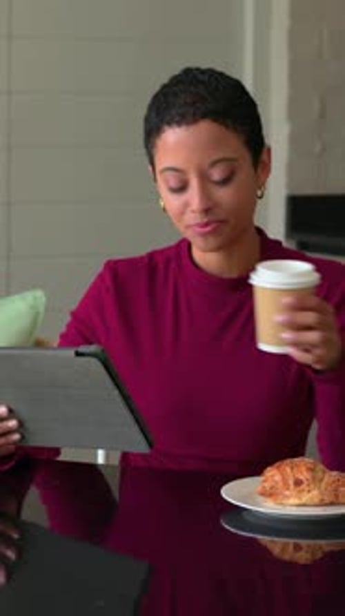 Vertical video: Reading tablet woman in burgundy top sipping cup, eating croissant smiling at home