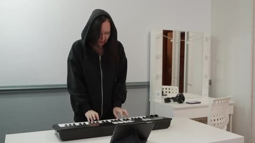 Female in Hoodie Practicing Keyboard Using Tablet in Lively Studio Scene