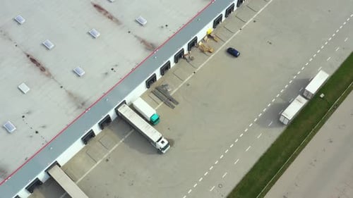 Aerial following shot of semi-trailer trucks travelling along a warehouse on the parking lot in the