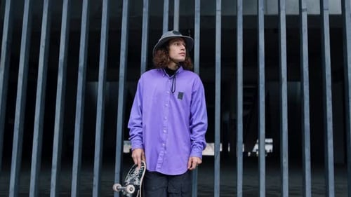 Stylish Cool Man Holding Skateboard Outdoor. Young Hipster Staying Near Gate With