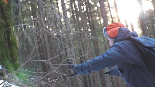 Profile of Young Hiker with Backpack Climbing Up on Snowy Hill and Keep Holding to Branches at Sunny
