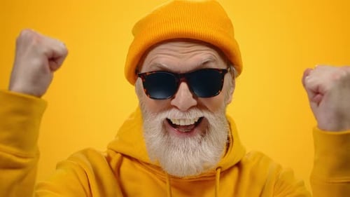 Joyful old man celebrates victory with clenched fists on yellow studio background