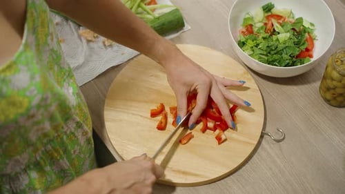 Close up of hands chopping red bell pepper into small pieces on a round wooden board beside a bowl