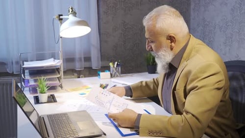 Man Works on Laptop and Reviews Documents in Office