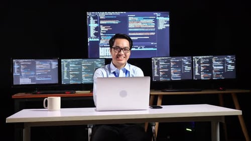 Programador sonriendo a la cámara y con los brazos cruzados mientras escribe código con una computadora portátil usando varios monitores