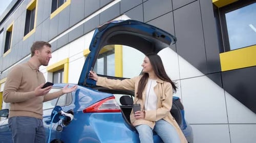 Happy young couple smiling and talking while drinking coffee standing outdoor at EV charging station