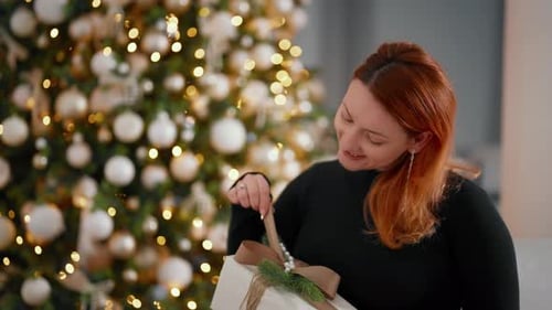 Woman Examining Wrapped Christmas Gift Near Holiday Tree