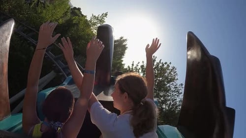 Two young girls with arms raised have fun riding adrenaline twisting roller coaster at amusement par