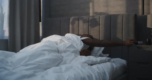 In Morning Handsome African American Man Wakes Up in Bed to Sound of Alarm Clock