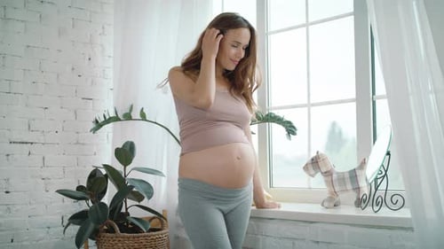 Happy Pregnant Woman Stroking Belly Near Window Indoors. Pretty Expectant Mother Standing
