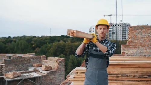 Walks with piece of board. Construction worker in uniform and safety equipment have job on building