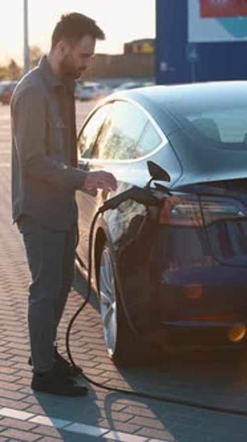 Man Charging Electric Car, Looking at Phone