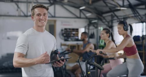 Fitness coach giving thumbs up and using a tablet while standing in a gym with his students