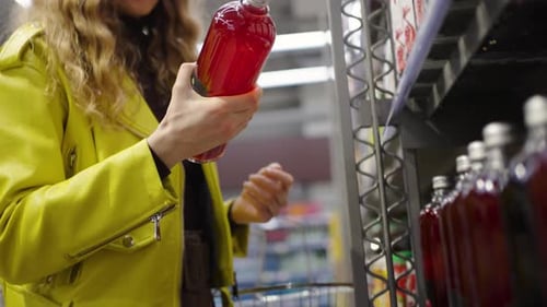Woman in a Vibrant Yellow Jacket Closely Examines a Bottle of Juice in the Supermarket Considering