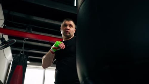 in a Boxing Fight Club Senior Muscular Trainer in a Black Tshirt and with Multicolored Bandages is