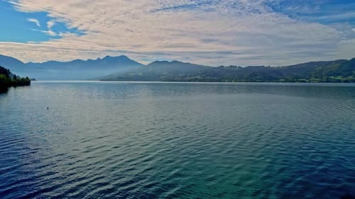 Aerial drone forward moving shot over a large lake surrounded by mountain range on a cloudy day.