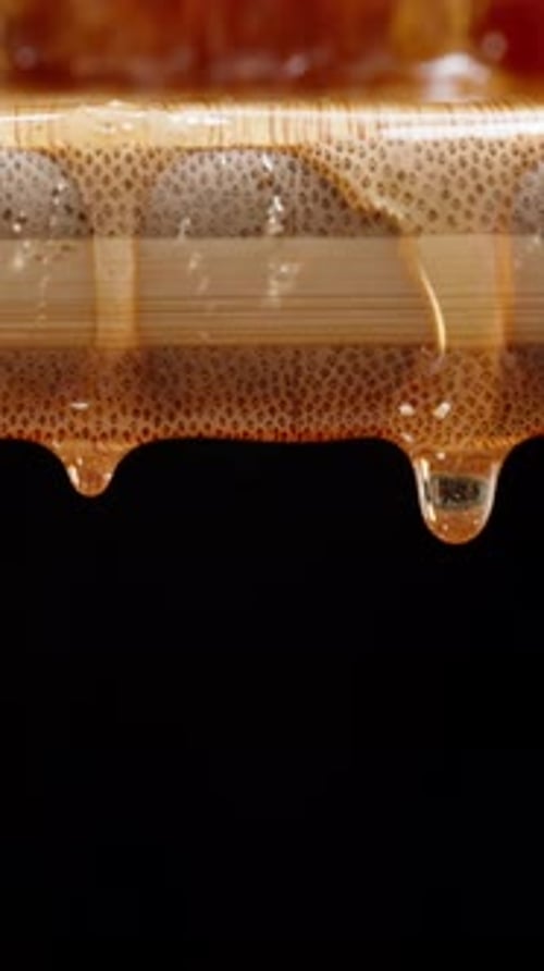 Vertical video. Drops of honey dripping from a wooden board onto a black background, close-up