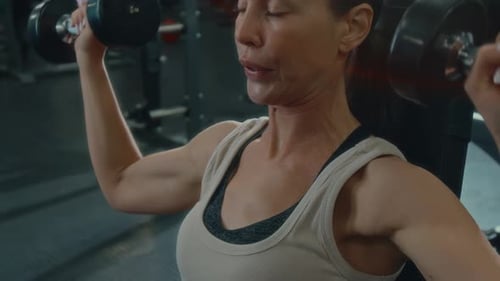 Woman Lifting Dumbbells while Performing Seated Shoulder Press in Gym