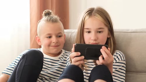 Hermano y hermana en edad escolar usando un dispositivo de teléfono inteligente relajándose juntos en el sofá de casa