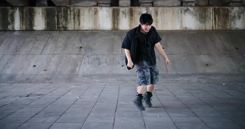 A stylish young man performs a freestyle dance under a footbridge in the city.