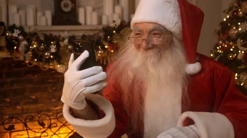 Santa Claus is Using the Phone Sitting at a Table Near a Christmas Tree and Decorated Fireplace