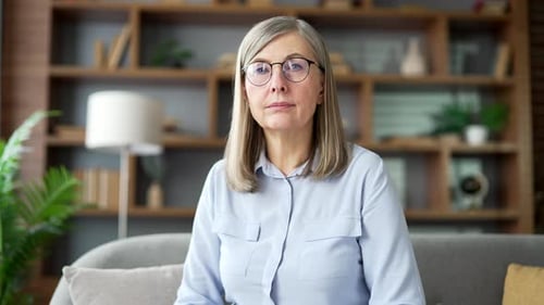 Webcam view. Senior gray haired female listens to online e-learning, video course looking at camera
