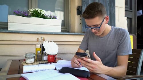 Young student focused on homework and online research at a city cafe table