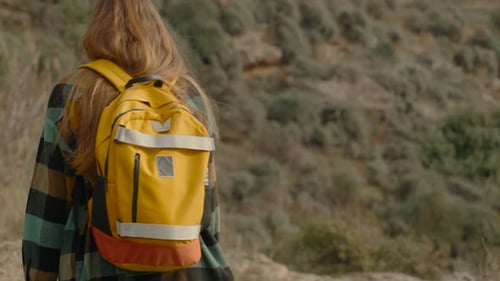 Young Female Traveler with a Bright Backpack Walking in a Mountain Valley, View from Behind.