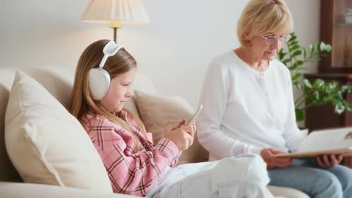 Girl Uses Phone Next to Grandmother Reading Book