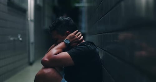 Man sitting in a narrow alley, head lowered, arms crossed over knees, overwhelmed by inner