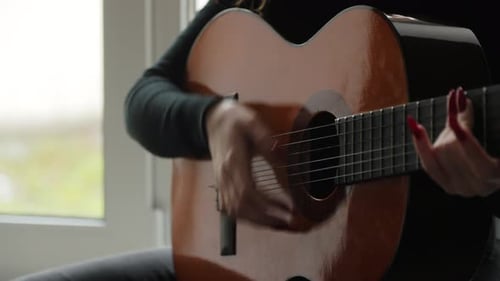 Close-up of a girl's hands playing chords on an acoustic guitar while sitting on a windowsill.