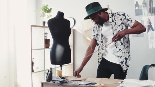 Stylish AfroAmerican Dressmaker in a Casual Look Designing Clothes Uses the Ruler for His Sketches