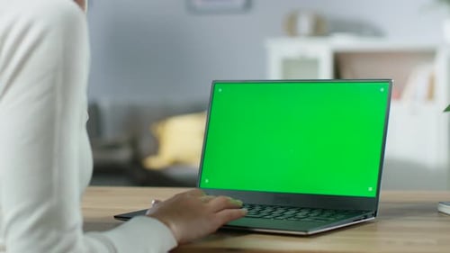 Over the Shoulder Shot of Woman's Hands Typing on a Green Mock-up Screen of the Laptop. In the Back