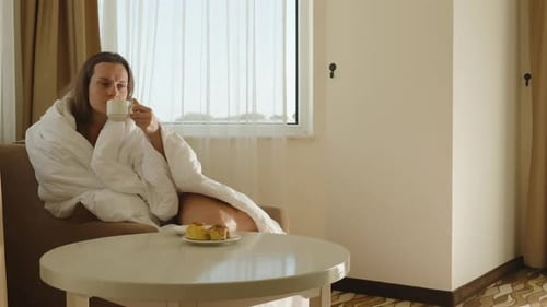 In the hotel room, a young woman sits on a chair wrapped in a blanket, drinking coffee while looking
