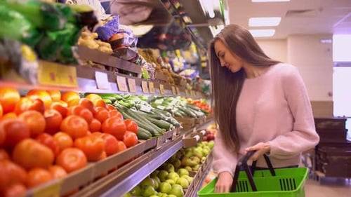 A Woman in a Supermarket on a Vegetable Shelf Buys Vegetables and Fruits Woman Chooses Cucumbers