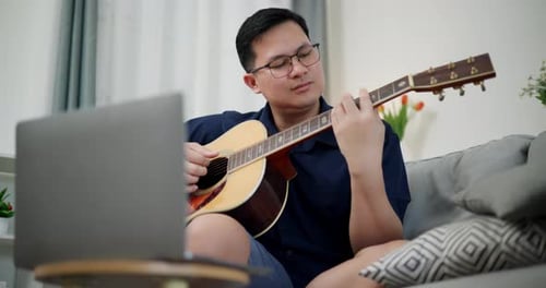 Man Plays Guitar at Home While Watching Laptop