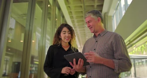 Man and woman smiling while holding tablet, collaborative team exchange in modern office hallway