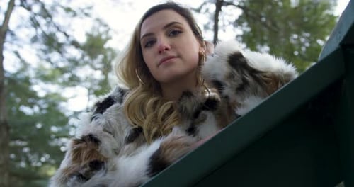 Élégante jeune femme blonde portant une veste de fourrure posant sur une balustrade, fond de forêt. Angle bas cl