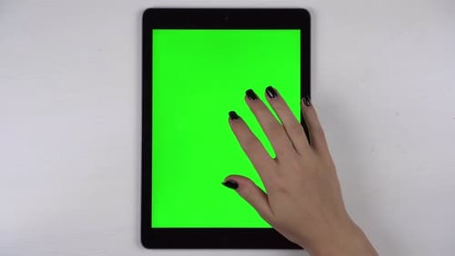 A Woman Taps on a Tablet Lying on a Table with a Green Screen