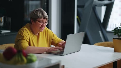 Senior Woman Working Remotely From Home Typing Text On Laptop Keyboard Sitting Alone At Kitchen