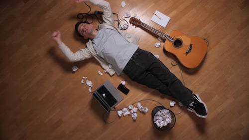 Young Male Musician Lying Exhausted on Floor Amid Crumpled Lyrics Laptop Microphone and Guitar