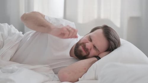 Bearded Man Waking Up in Bed Rubbing Eyes